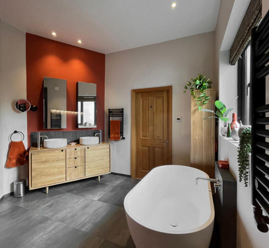 Photo of a bathroom installation in Wilmslow, Cheshire. shows a freestanding white resin bath, black wall mounted towel radiators, dark grey porcelain flor tiles and a oak vanity unit